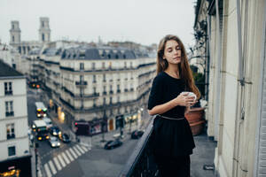 Sensual pretty woman with eyes closed standing on balcony with cup in Paris, France.. Sensual woman on balcony - INGF12419