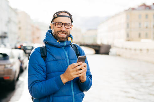 Happy man with beard wearing eyeglasses dressed in blue anorak holding backpack and mobile having happy look travelling in city watching sightseeings and making photos having exursion outdoor - INGF12420