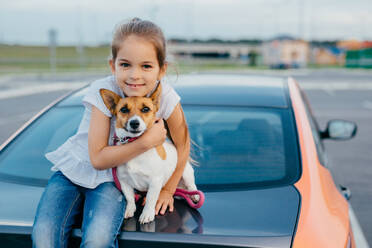 Little girl with appealing appearance, hugs her favourite pet, have journey together with parents by car, sit at trunk, pose for making photo. Children, animals, rest and transportation concept - INGF12422