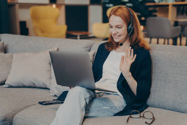 Young woman using laptop and wireless headset for internet video call with friends, waving hand in greeting gesture, broadly smiling while looking on computer screen. Online communications concept. Young woman using laptop and headset for internet video call waving hand in greeting gesture - INGF12425