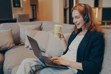 Aufgeregte rothaarige lächelnde Frau mit drahtlosem Headset und Laptop vor sich, die ein Online-Gespräch mit Kollegen führt, während sie in entspannter Atmosphäre auf dem Sofa im Café sitzt und eine Tasse Kaffee in der Hand hält. Aufgeregte rothaarige lächelnde Frau mit drahtlosem Headset und Laptop, die auf dem Sofa im Café sitzt - INGF12426