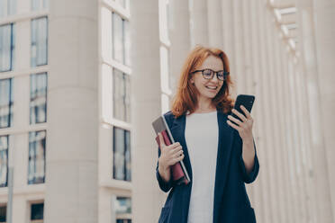 Happy european female holding mobile phone and smiling, reading good news from business partner while standing outside near office building with laptop, chatting with friend during coffee break. Happy european female holding mobile phone and smiling, reading good news from business partner - INGF12428