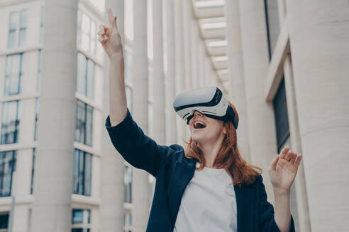 Augmented reality in business. Young excited happy female enterpruer wearing portable VR goggles trying to touch something in virtual reality with her finger, standing alone on city street. Excited female enterpruer wearing portable VR goggles trying to touch something in virtual reality - INGF12429