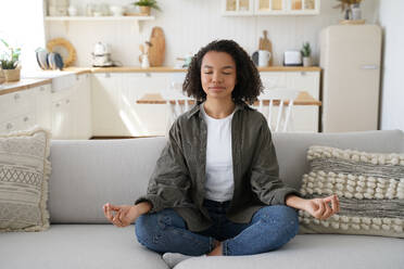 Teenage afro girl is practicing yoga at cosy home. Young woman sitting on couch in lotus pose. Concentration and balance. Wellness and emotional wellbeing, tranquility and recreation, stress relief.. Teenage afro girl is practicing yoga at cosy home. Wellness and emotional wellbeing, stress relief. - INGF12431