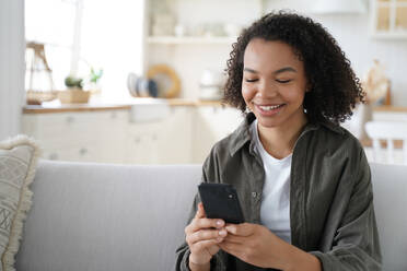 Happy african american teen girl share good news on social network via mobile phone sitting on sofa at home. Smiling mixed race young female enjoying shopping online using smartphone apps.. Happy african american teen girl holds smartphone using social network apps, shopping online at home - INGF12432