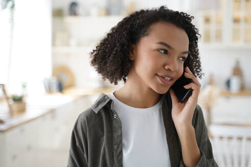 Modern african american girl talking on smartphone at home, stylish young woman with afro hairs answer mobile phone call, having conversation with friends or relatives indoors.. Modern african american young girl talking on smartphone, answering mobile phone call at home - INGF12433