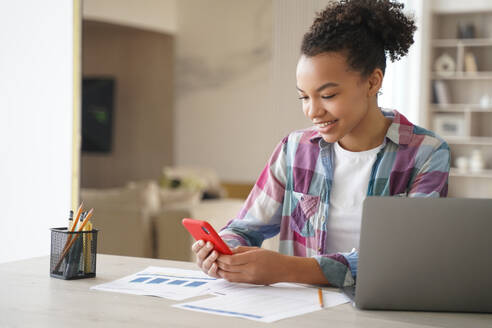 Afro teenage girl is distracting while studying remote. Teenage girl sitting in front of laptop, clicks mobile phone and chatting. African american student gets remote education at school or college.. Afro teenage girl is distracting while studying remote. Student clicks mobile phone and chatting. - INGF12440