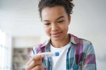 Young biracial girl using digital thermometer happy with her body temperature. Smiling african american woman checking temp before trip during coronavirus covid pandemic.. Young biracial girl uses digital thermometer happy with body temperature during coronavirus pandemic - INGF12444