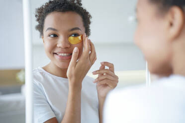 Mirror reflection of young hispanic lady. Happy afro girl is applying eye patches to her face. Skin moisturizer with collagen serum. Anti age patches. Morning skincare. Beauty routine of teenage girl.. Mirror reflection of young hispanic lady with eye patches. Skin moisturizer with collagen serum. - INGF12446