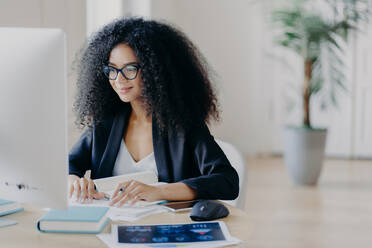 Freelance Afro woman works remotely, writes information, focused at computer screen with delighted expression, poses at workplace in spacious cabinet, potted plant in background. E learning concept - INGF12448
