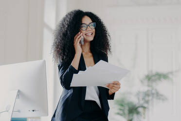 Pleased successful businesswoman with Afro hairstyle wears spectacles to provide eyes protection, holds documents, makes telephone call to business partner, stands in cabinet near workplace. - INGF12451