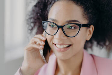 Headshot of beautiful smiling dark skinned woman wears optical glasses, holds modern cell phone, has well cared complexion, talks about future investments with business partner, looks aside. - INGF12454