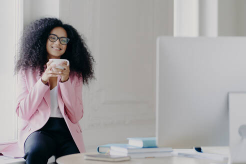 Joyful curly haired female freelancer or blogger has coffee break, holds white mug with beverage, poses near window, focused in computer, watches useful training video, wears near formal clothes - INGF12455