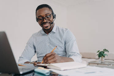 African american man wearing headset working distantly on computer at home office. Black guy student in headphones learns online at laptop, takes notes. E-learning, distance education.. African american man in headset learns online at laptop, takes notes. Elearning, distance education - INGF12457