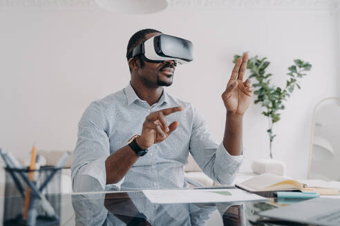 Businessman in vr headset clicks virtual buttons. Freelancer is working on design project. African american man in futuristic vr headset at home. Modern digital technology for business and creativity.. Businessman in vr headset clicks virtual buttons. Modern technology for business and creativity. - INGF12460