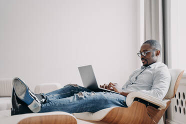 Focused african american man freelancer working online at laptop, typing email or message, sitting in modern comfortable armchair. Black male businessperson developing business project. Remote work. African american man working online on business project at laptop, sitting in armchair. Remote job - INGF12462