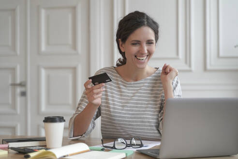 Young happy Spanish woman holding credit card and feeling excited while doing online shopping on laptop at home, looking at computer screen and purchasing goods or services in internet store. Young happy Spanish woman hold credit card and feeling excited while doing online shopping on laptop - INGF12464