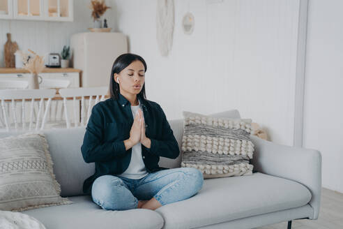 Calm young woman deep breathing practicing yoga sitting on couch at home. Female meditates folded hands in namaste gesture in lotus posture on sofa. Wellness, stress relief concept.. Calm woman breathing practicing yoga sitting in lotus pose on couch at home. Wellness, stress relief - INGF12468