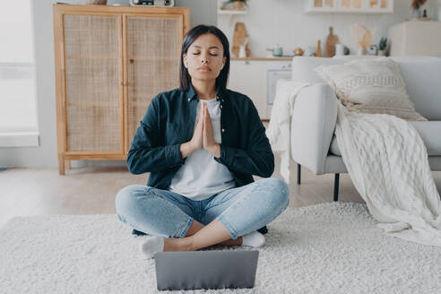 Frau meditiert am Laptop während einer Online-Yoga-Stunde, sitzend in Lotus-Asana auf dem Boden zu Hause. Ruhige Frau hört Audio-Affirmationen fühlt Gelassenheit. Wellness, Ruhe, Stressabbau Konzept. Frau übt Yoga, meditiert am Laptop in Lotus-Asana auf dem Boden zu Hause. Wellness, Stressabbau - INGF12469