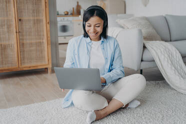 Lächelnde Frau mit Kopfhörern, die an einem Laptop arbeitet und von zu Hause aus online lernt, auf dem warmen Boden sitzend. Freundliche Studentin mit drahtlosem Headset, die ein Webinar hört. Fernunterricht, E-Learning. Lächelnde Frau mit Headset, die an einem Laptop arbeitet und von zu Hause aus online lernt, auf dem Boden sitzend. E-Learning - INGF12471