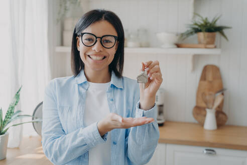 Smiling positive spanish woman is showing house key in her hand. Success and achievement concept. Happy real estate owner. Mortgage loan and investment. Modern scandinavian interior of kitchen.. Smiling positive spanish woman is showing house key in her hand. Happy real estate owner. - INGF12472