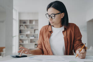 Focused woman in glasses manage household budget calculate expenses for saving money. Serious female work with financial papers, count rent, mortgage fees, sitting at desk at home.. Serious woman in glasses manage household budget count financial expenses for saving money at home - INGF12474