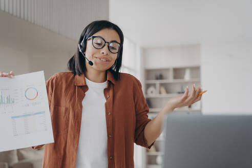 Businesswoman wearing glasses, headset, talking by corporate video call showing infographic. Professional female business coach holding document with graphs, discussing project online.. Businesswoman in glasses, headset, discuss business project online by video call showing infographic - INGF12476