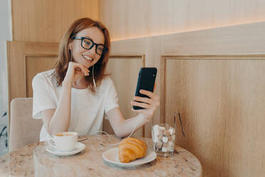 Beautiful redhead woman in earphones sitting in cafe and making video call on mobile phone, talking with friends or followers during video chat on smartphone, blogger enjoying breakfast in coffee shop. Beautiful redhead woman in earphones sitting in cafe and making video call on mobile phone - INGF12477