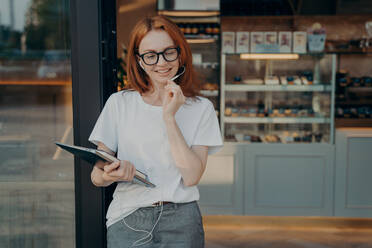 Positive redhead woman uses earphones with built in microphone tablet to communicate with friends and colleagues has conversation via internet wears spectacles white t shirt grey formal trousers. Positive redhead woman uses earphones with built in microphone tablet to communicate - INGF12478