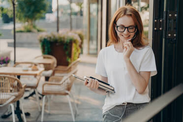 Happy beautiful redhead young woman makes distant call uses earphones with microphone carries modern gadgets poses outdoor at street against cafe blurred background. People and lifestyle concept. Beautiful redhead young woman makes distant call uses earphones with microphone - INGF12480