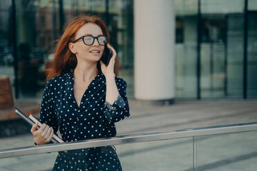 Thoughtful redhead woman wears spectacles polka dot dress has telephone conversation looks into distance stands near blurred office background carries necessary things for working. Lifestyle. Thoughtful redhead woman wears spectacles polka dot dress has telephone conversation - INGF12482