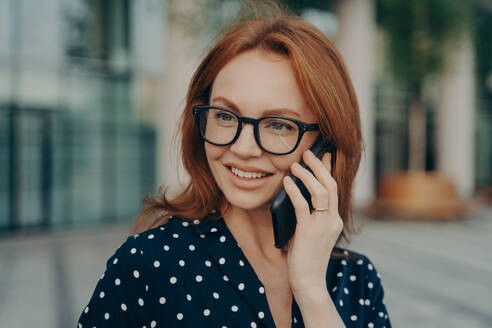 Photo of beautiful redhead woman has telephone talk uses smartphone for call looks away wears spectacles polka dot clothes poses against blurred background outdoor phones via mobile application. Beautiful redhead woman has telephone talk uses smartphone for call - INGF12483