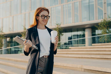 Beautiful stylish redhead businesswoman with laptop drinking coffee from disposable paper cup while going to work in morning, female business professional walking outdoors on city street and smiling. Stylish businesswoman with laptop drinking coffee from disposable paper cup while going at work - INGF12484