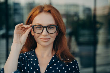 Portrait of confident beautiful red-haired business woman adjusting spectacles eyeglasses while posing on city street in summer, looking at camera with confidence, blurred urban background. Confident beautiful red-haired business woman adjusting spectacles eyeglasses posing on street - INGF12485