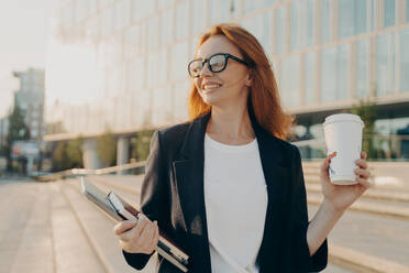 Overjoyed happy ginger woman in spectacles drinking coffee and holding laptop while walking outdoors on sunny day, business woman enjoying her way to office in morning, smiling and looking asidee. Overjoyed happy ginger woman in spectacles drinking coffee and holding laptop while walking outdoors - INGF12486
