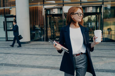 Beautiful redhead female entrepreneur standing outdoors with coffee cup and laptop in hands, leaving office, businesswoman in spectacles dressed in formal clothes waiting for taxi and looking aside. Beautiful redhead female entrepreneur standing outdoors with coffee cup and laptop in hands - INGF12488