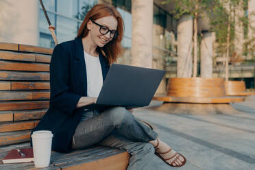 Happy cheerful european ginger woman working remotely on laptop outside, sitting on wooden bench and drinking morning coffee, female in eyeglasses enjoying remote work outdoors, checking e-mail. Happy cheerful european ginger woman working remotely on laptop outside, sitting on wooden bench - INGF12489