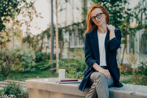 Frustrated redhead young woman feels very tired suffers from neck pain sits in park outdoors wears transparent glasses and formal wear focused down thinks about something fatigue after work.. Frustrated redhead young woman feels very tired suffers from neck pain sits in park - INGF12491