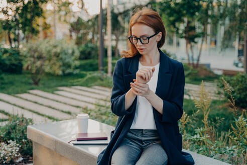 Young tired redhead businesswoman measuring pulse on wrist, checking heart rate after stressful working day in office, overworked female sitting in park and relaxing, feeling exhausted. Healthcare. Young tired redhead business woman measuring pulse on wrist, checking heart rate, sitting in park - INGF12492