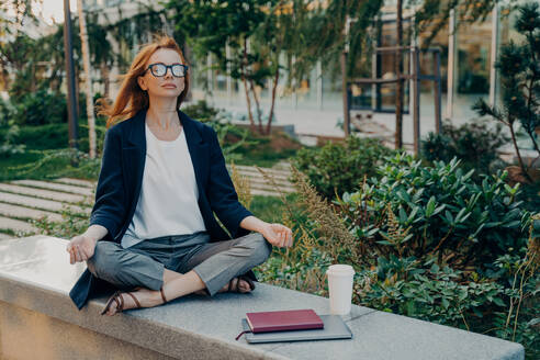 Peaceful redhead business woman dressed formally holding hands in mudra gesture, sitting in lotus pose on concrete bench and meditating with closed eyes, relaxing after remote work on laptop in park. Peaceful business woman holding hands in mudra gesture, sitting in lotus pose and meditating in park - INGF12493