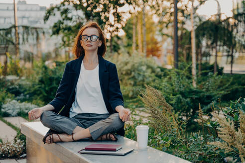 Full length shot of relaxed ginger woman meditates sits in lotus pose wears formal clothes keeps legs crossed breathes deeply spends free time in park surrounded by takeaway coffee tablet and notepad. Full length shot of relaxed ginger woman meditates sits in lotus pose wears formal clothes - INGF12495