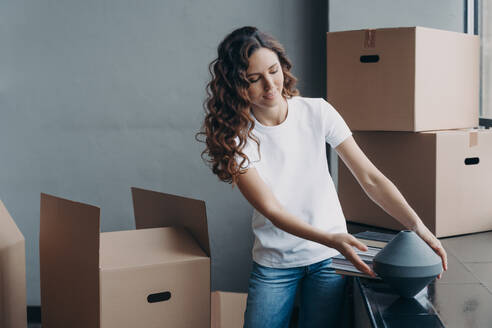 Happy girl unpacking cardboard boxes and taking the vase. Smiling attractive hispanic woman packing boxes to move. Relocation to new apartment. Mortgage and real estate concept.. Happy girl unpacking cardboard boxes. Relocation to new apartment. Mortgage and real estate concept. - INGF12496