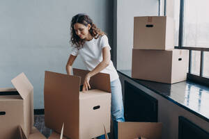 Happy young european woman unpacking boxes. Girl near window in white t-shirt indoors. Woman is opening cardboard box and smiling. Relocation and independence concept.. Happy young european woman unpacking boxes. Girl near window opening cardboard box and smiling. - INGF12497
