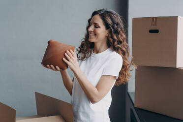 Relocation and delivery service concept. Happy girl unpacking cardboard boxes and holding the vase. Woman in casual outfit. Smiling attractive spanish woman packing boxes to move.. Relocation and delivery service concept. Happy girl unpacking cardboard boxes and holding the vase. - INGF12498