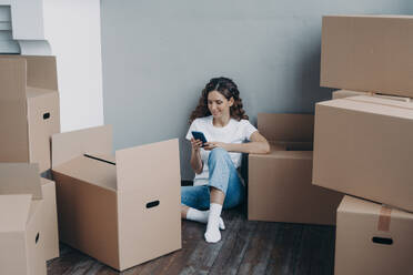 Happy young european woman with smartphone is packing boxes. Girl is chatting on mobile phone and sitting on the floor. Easy moving and shipping service order concept.. Happy young european woman with smartphone is packing boxes. Shipping service order concept. - INGF12500