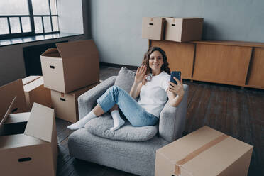 Excited young woman unpacking things at new place. Lady having video call on phone in new apartment. Successful independent european girl shows her dream house. Mortgage loan concept.. Excited young woman unpacking things at new place. Lady having video call on phone in new apartment. - INGF12503