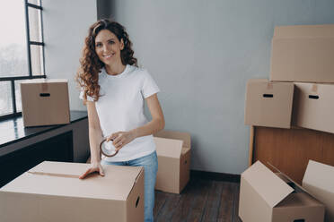 Mover is packing cargo with duct tape. Young lady is wrapping cardboard boxes with packing tape. Happy hispanic girl relocates. Moving service worker preparing boxes for shipment.. Mover is packing cargo with duct tape. Young lady is wrapping cardboard boxes with packing tape. - INGF12508