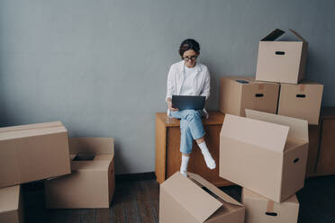 Spanish business lady rents apartment. Young businesswoman moves. Happy girl is sitting among carton boxes and working on laptop. Mortgage loan and real estate purchase.. Spanish business lady rents apartment. Happy girl is sitting among boxes and working on laptop. - INGF12510