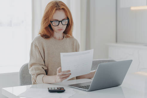 Young focused red-haired woman in spectacles calculating budget at home, sitting at table with laptop, holding paper taxes bills and reading financial information, female managing family budget. Young focused red-haired woman sitting at table with laptop and holding paper taxes bills - INGF12513