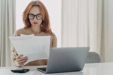 Serious redhead Caucasian woman calculates bills holds paper documents has focused look uses laptop computer and calculator thinks how to save money poses at workplace against home interior.. Serious redhead Caucasian woman calculates bills holds paper documents has focused look - INGF12514